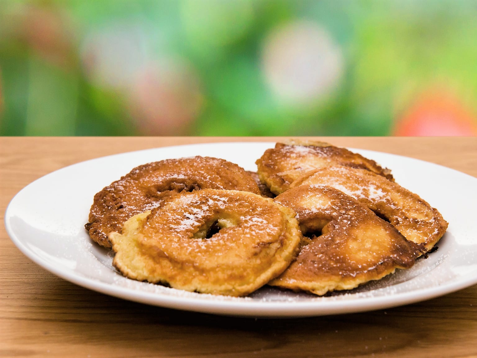 Four golden Apfelküchle rings on a cream plate, one broken open to show the pale apple ring inside the blistered batter, cinnamon sugar clinging to the crust, a jug of Vanillesoße alongside in natural light