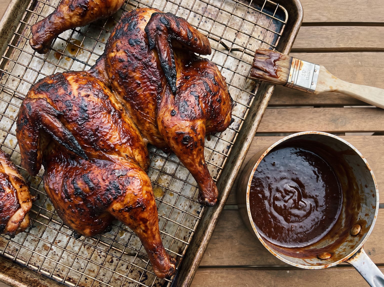 Barbecue chicken pieces on a rack with mahogany lacquered skin and darker charred edges, with a brush and a pot of sauce