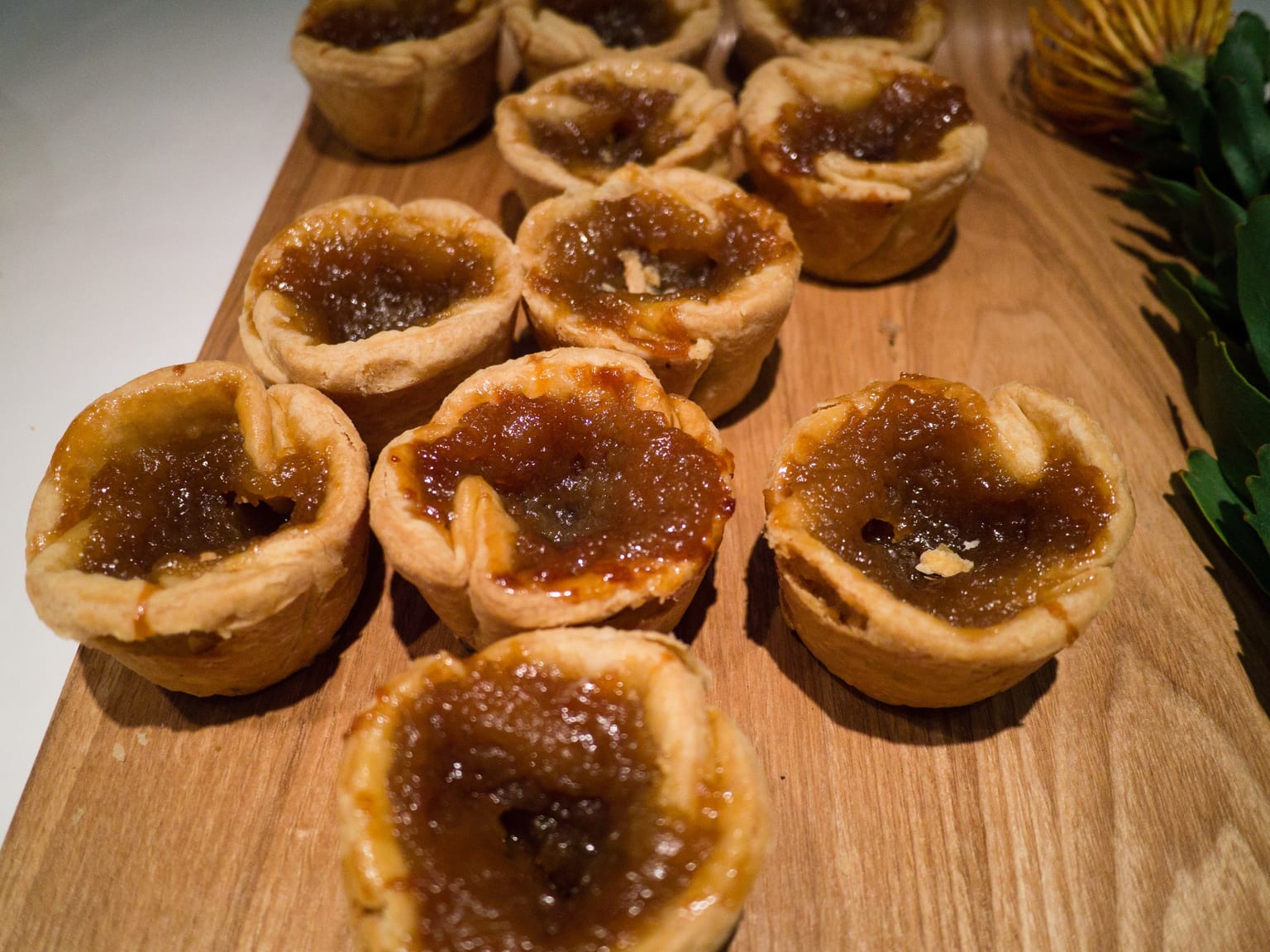 A dozen butter tarts cooling on a wire rack on a cream linen cloth, tops crazed and dark amber, one broken open on a plate to show the filling running out over the pastry