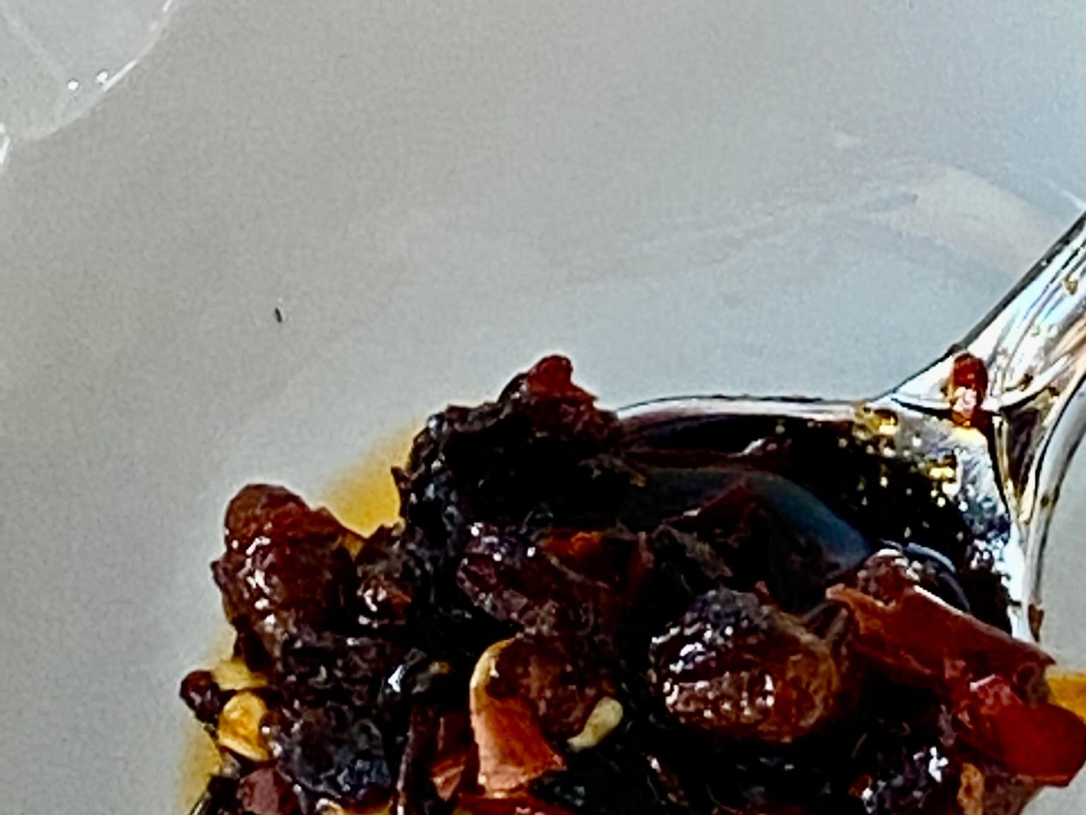 A glass jar of chili crisp on a cream surface in natural light, deep red oil above a thick layer of chile flakes, peanuts, and fried shallot, a spoon resting alongside