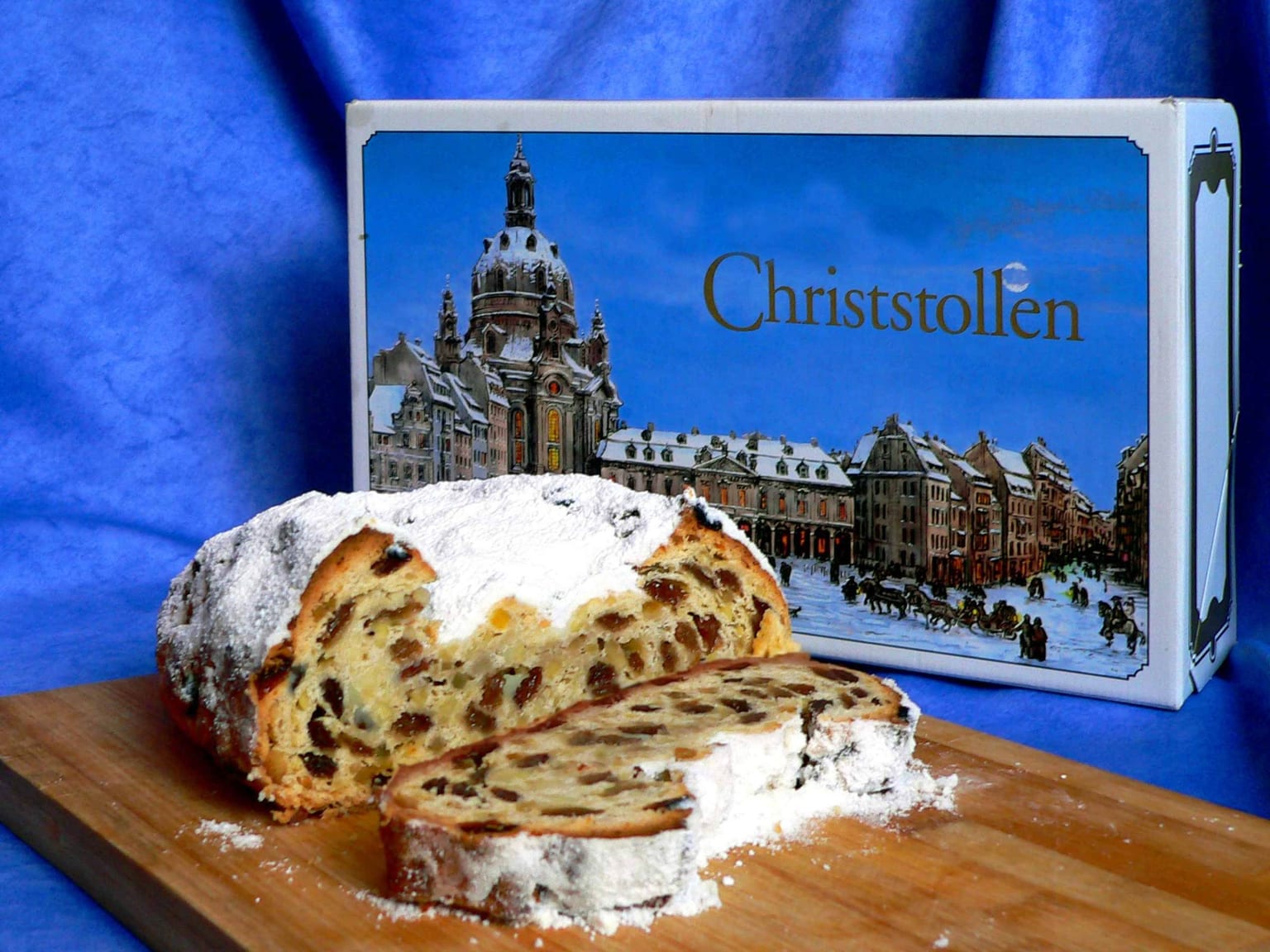 A Christstollen under a thick white blanket of powdered sugar on a wooden board in soft natural light, two slices cut to show the fruit and a marzipan core, cream linen and a small knife alongside