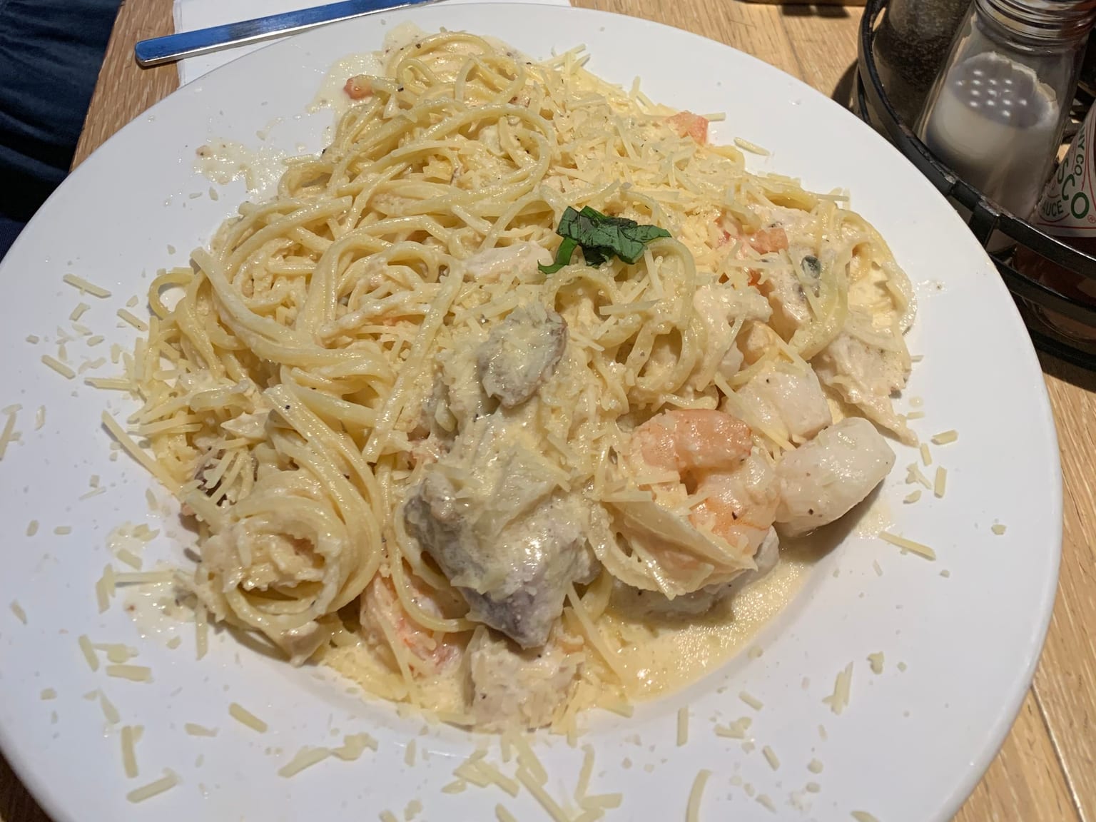 A wide plate of spaghetti in a pale cream sauce with prawns, chunks of white fish and mussels, heavily scattered with grated parmesan and a single basil leaf