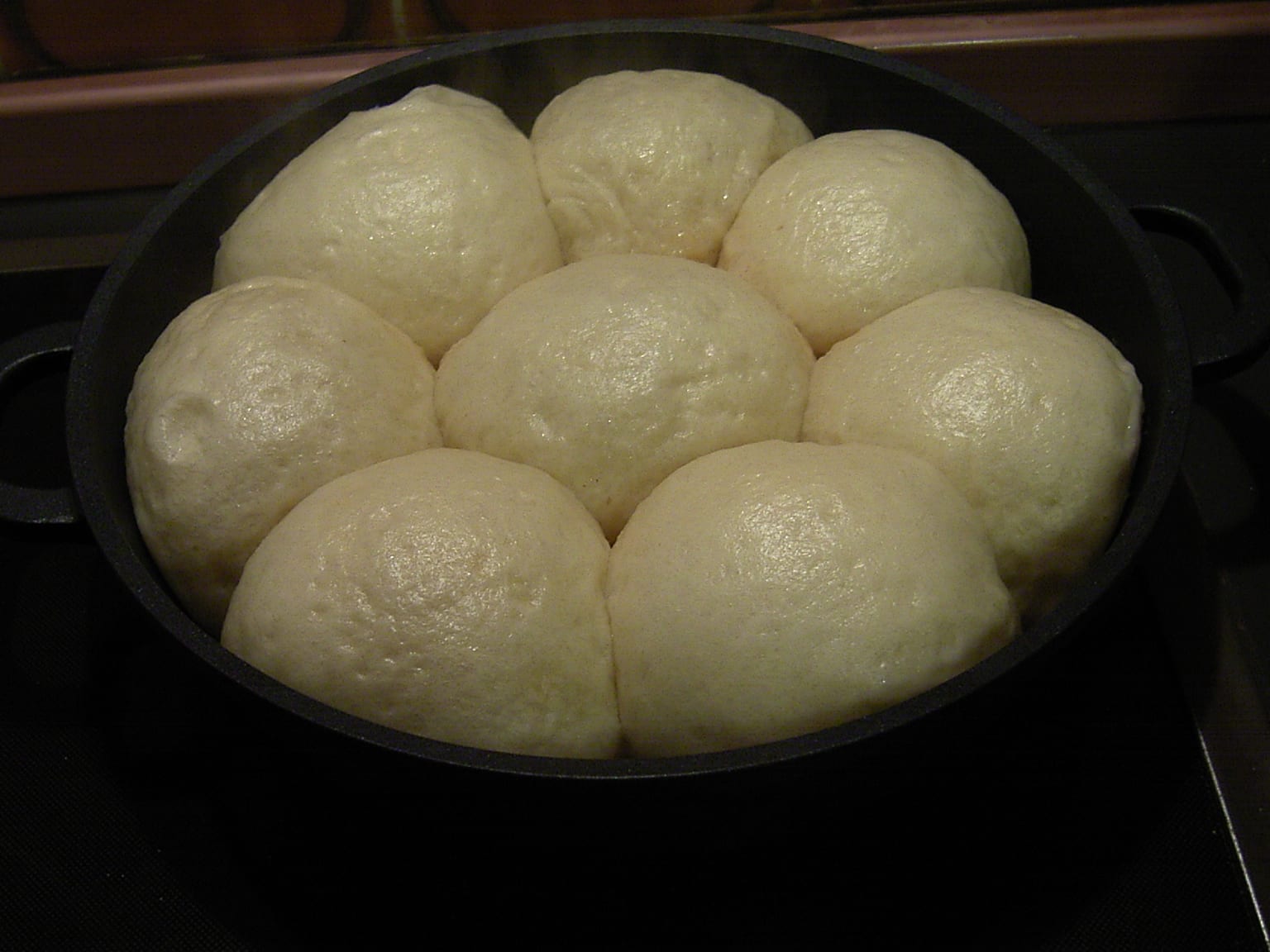 Four Dampfnudeln lifted from a wide skillet onto a cream plate, pale domed tops and dark caramelised crusts underneath, Vanillesoße poured around them in natural light