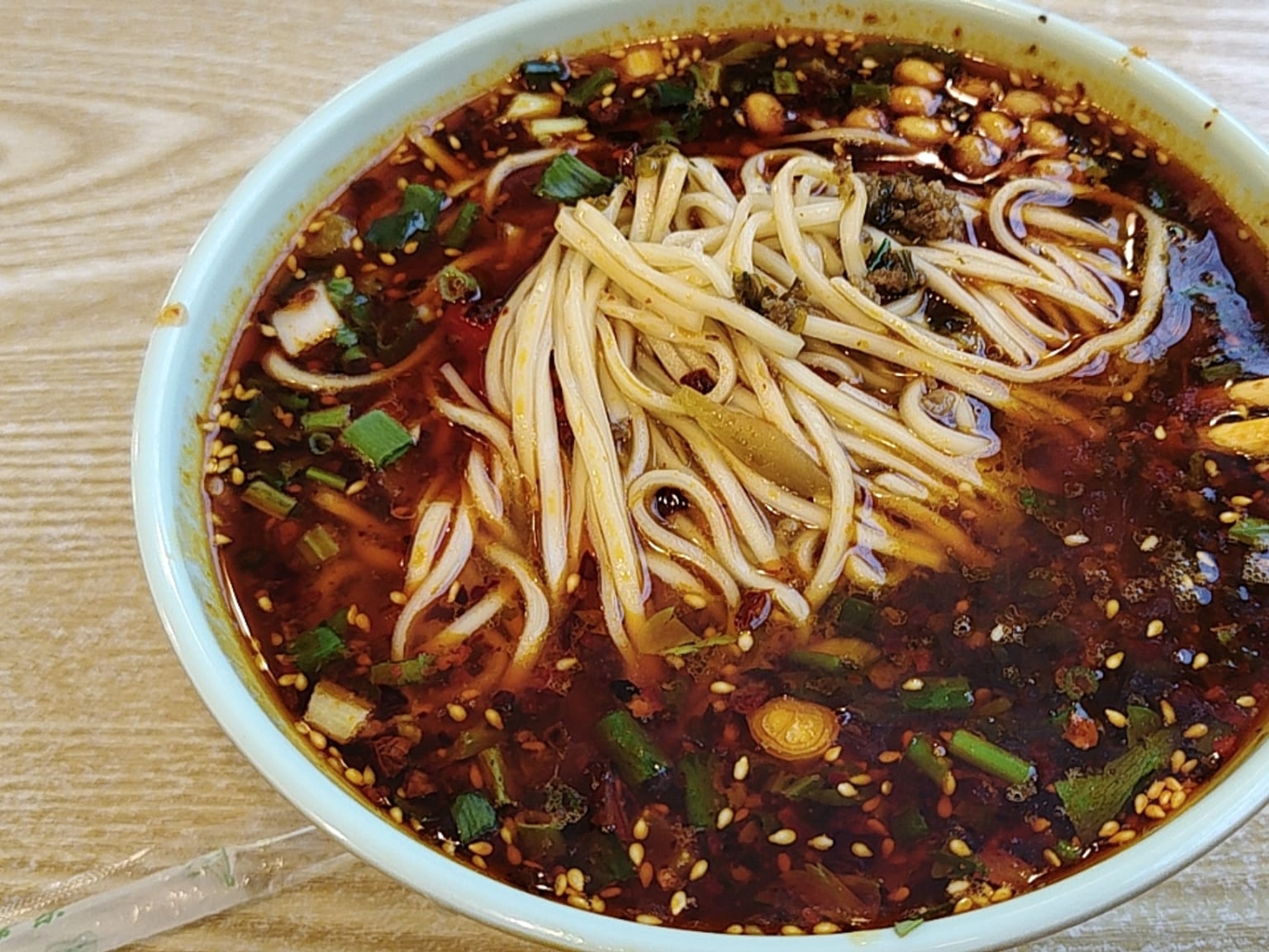 A small cream bowl of dan dan noodles, dark red chili oil at the bottom, thin wheat noodles, crumbled dark pork and preserved mustard green on top, crushed peanuts and scallions scattered over