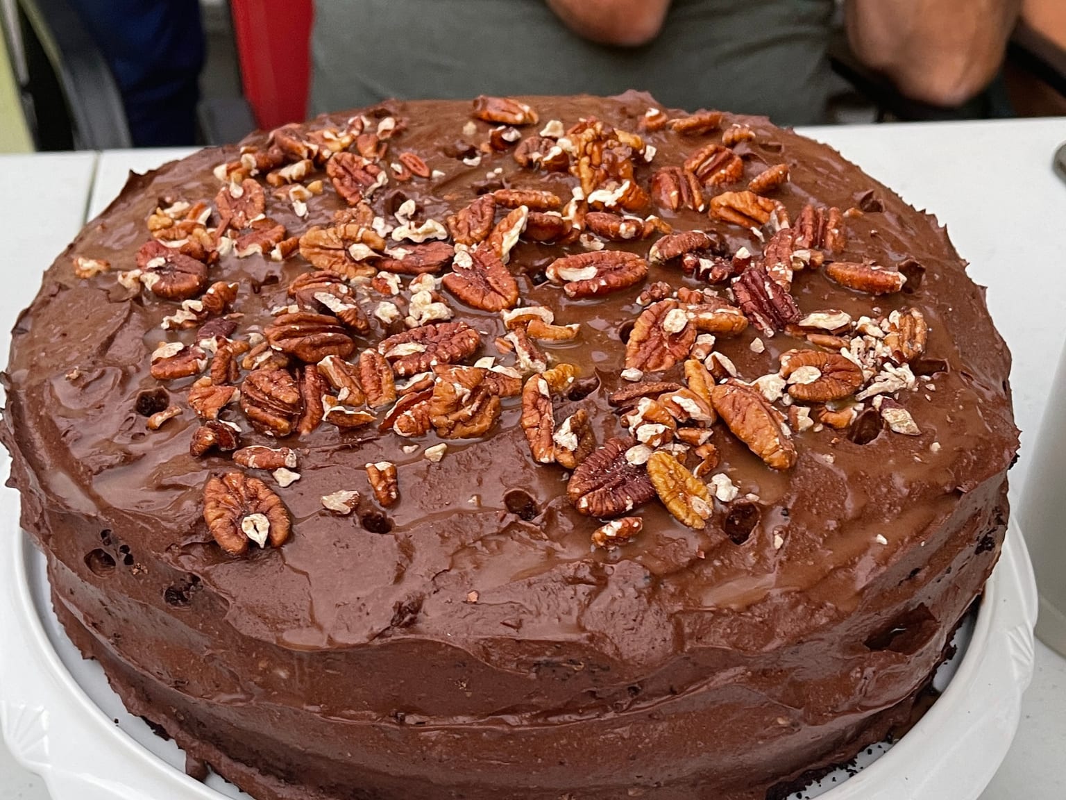 A two-layer chocolate cake covered in dark chocolate frosting on a white stand, the top scattered with toasted pecan halves and glossy caramel