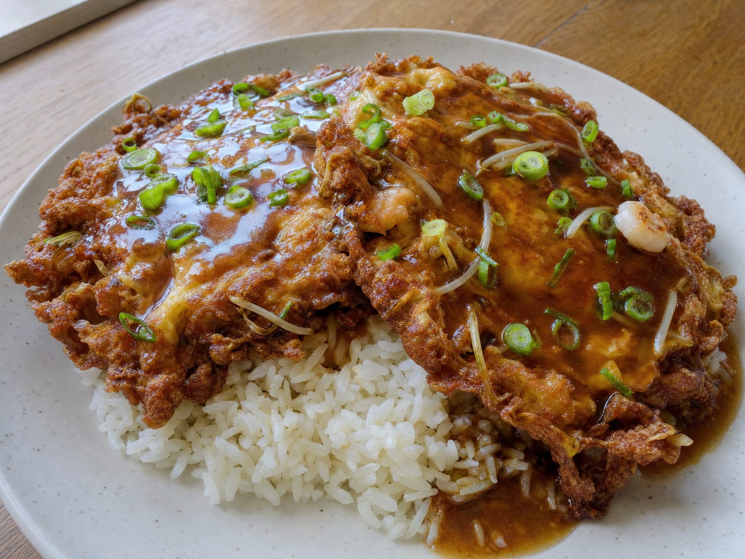 Two golden egg foo yong patties with ruffled edges under a glossy brown gravy, scattered with scallions