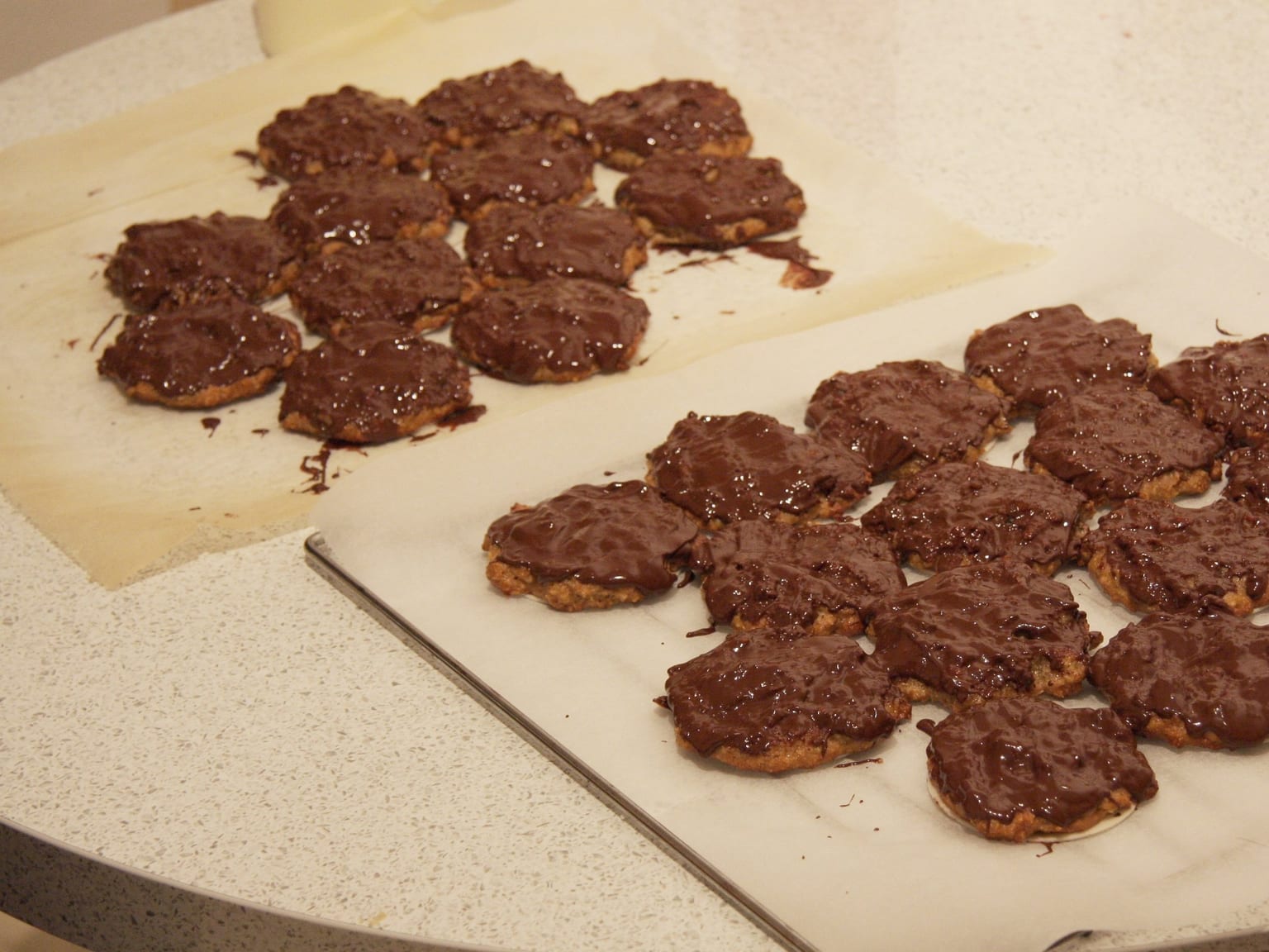 Round Elisenlebkuchen on their wafers cooling on a rack, half glazed in dark chocolate and half in white sugar glaze, with candied peel, whole almonds, and a small bowl of spice mix on cream linen