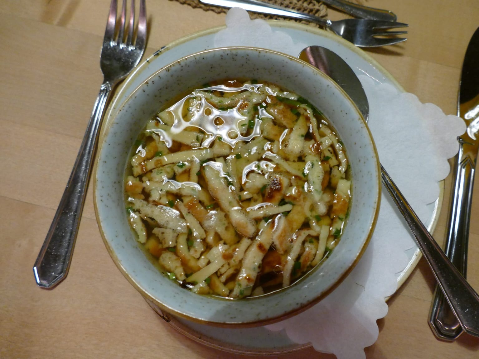 A shallow cream bowl of clear beef broth with pale golden pancake ribbons and sliced chives, natural light from the side