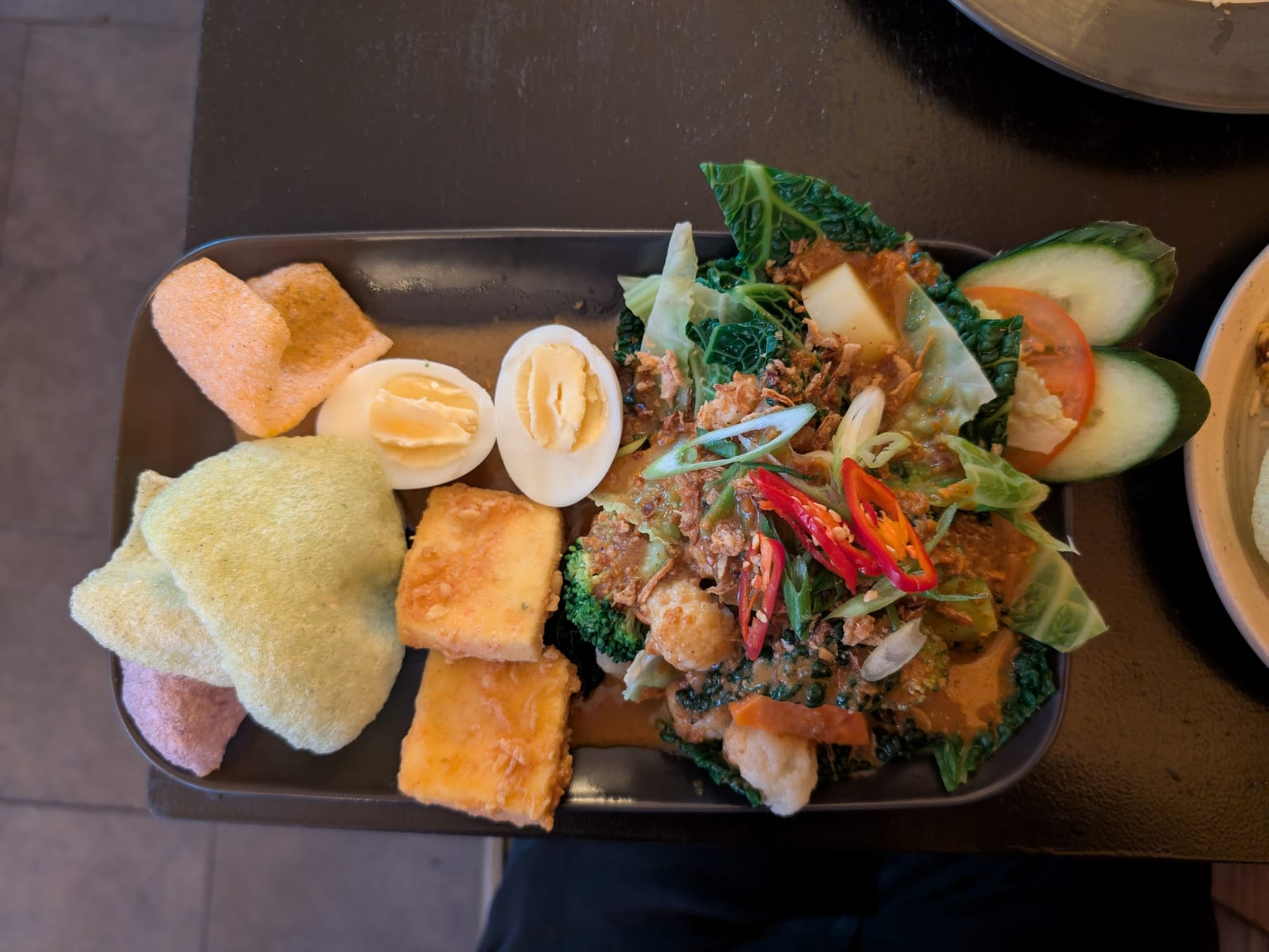 A large cream platter of gado-gado, blanched green beans, cabbage, bean sprouts and spinach arranged in sections with potato slices, halved boiled eggs, golden fried tofu and tempeh, cucumber, thick brown peanut sauce spooned over and fried shallots on top