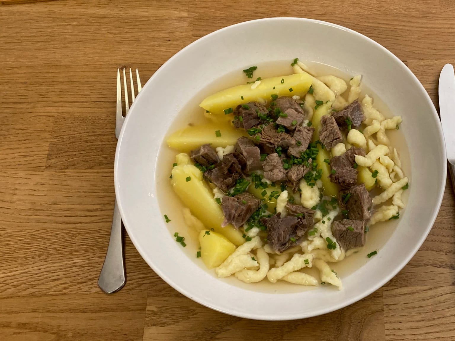 A deep cream bowl of clear broth with diced beef, potato cubes, and Spätzle, topped with dark golden fried onions and parsley, natural light from the side
