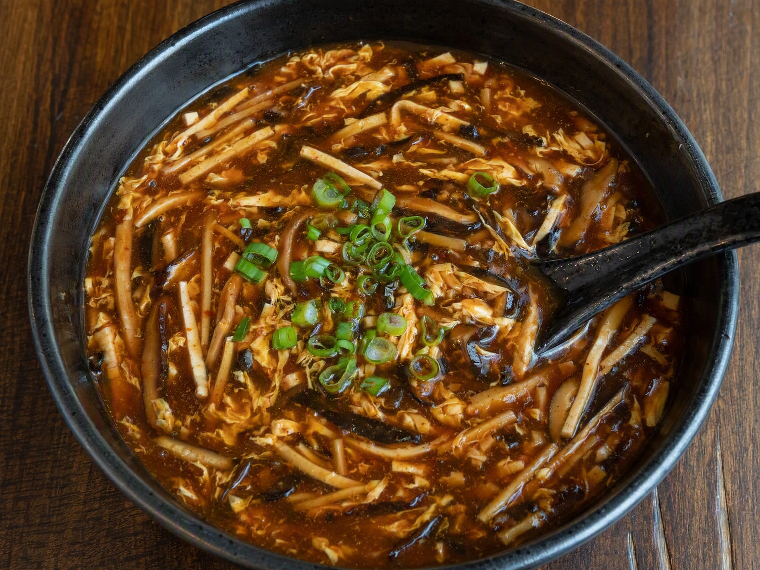 A dark glossy bowl of hot and sour soup with egg ribbons, tofu and mushroom strips, scattered with scallions