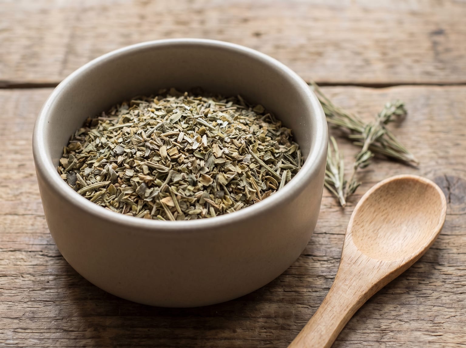 A small bowl of dried Italian herb blend with separate green-grey flakes of oregano, basil, rosemary and thyme visible, with a spoon and dried sprigs beside it