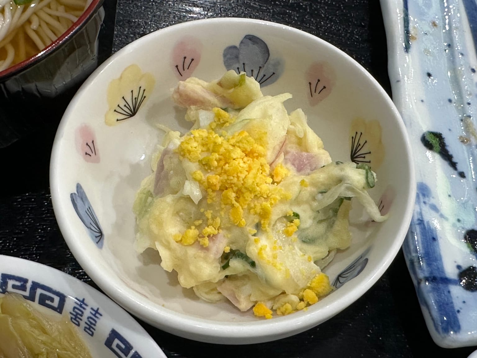 Japanese potato salad in a pale ceramic bowl on a cream cloth, lumpy and pale yellow, flecked with orange carrot, green cucumber, and chopped egg