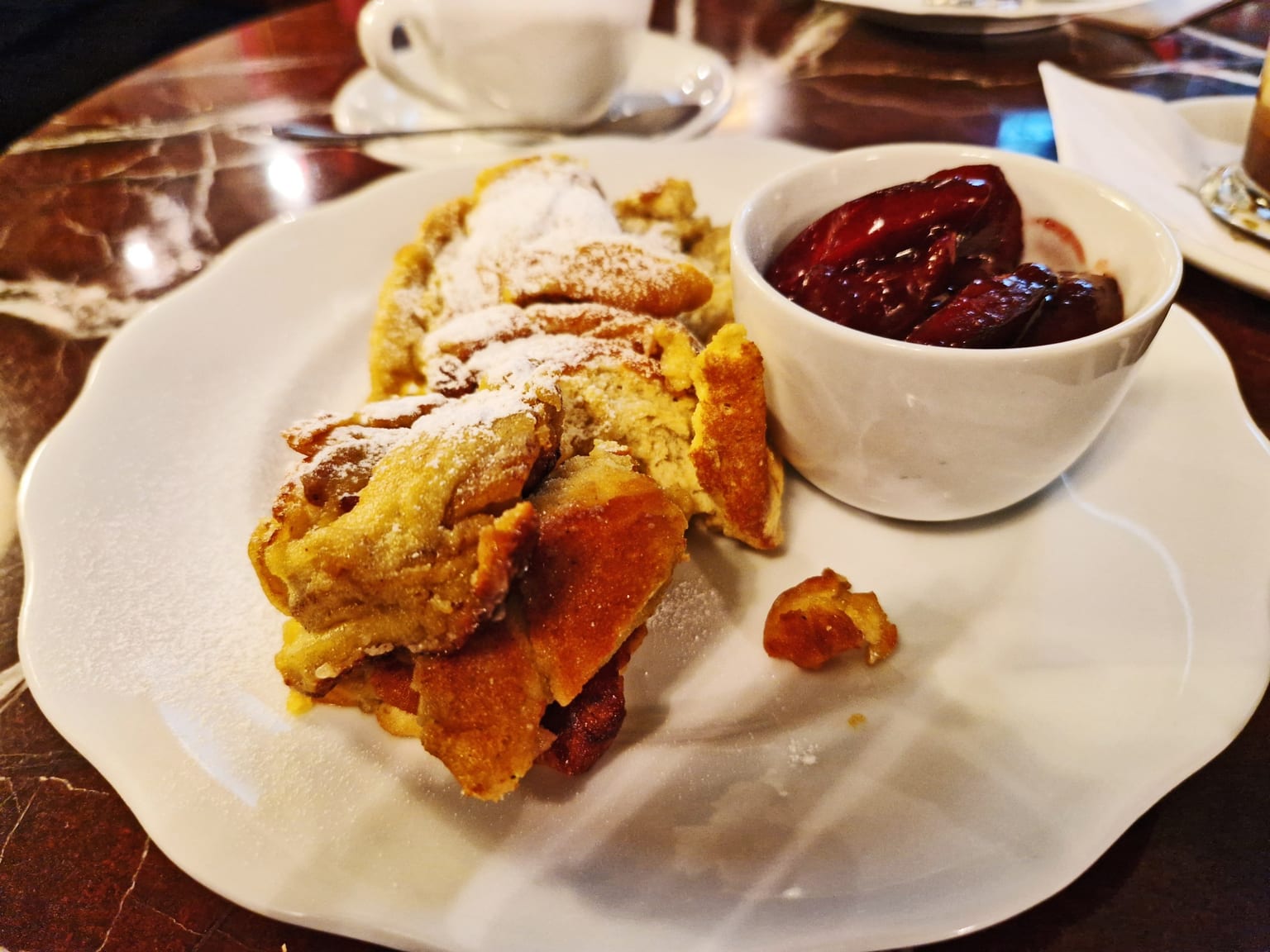 A cast iron skillet of torn Kaiserschmarrn pieces with caramelised golden edges under a heavy dusting of icing sugar, a small bowl of dark red Zwetschgenröster beside it on a cream cloth in natural light
