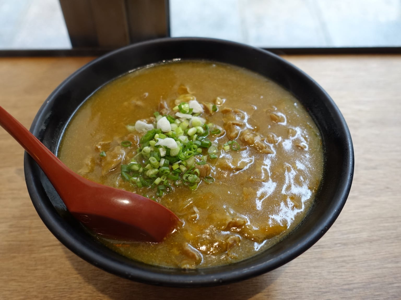 A deep cream bowl of curry udon in natural light, glossy pale brown curry broth clinging to thick white noodles, sliced pork and a halved soft egg on top, green spring onion scattered over