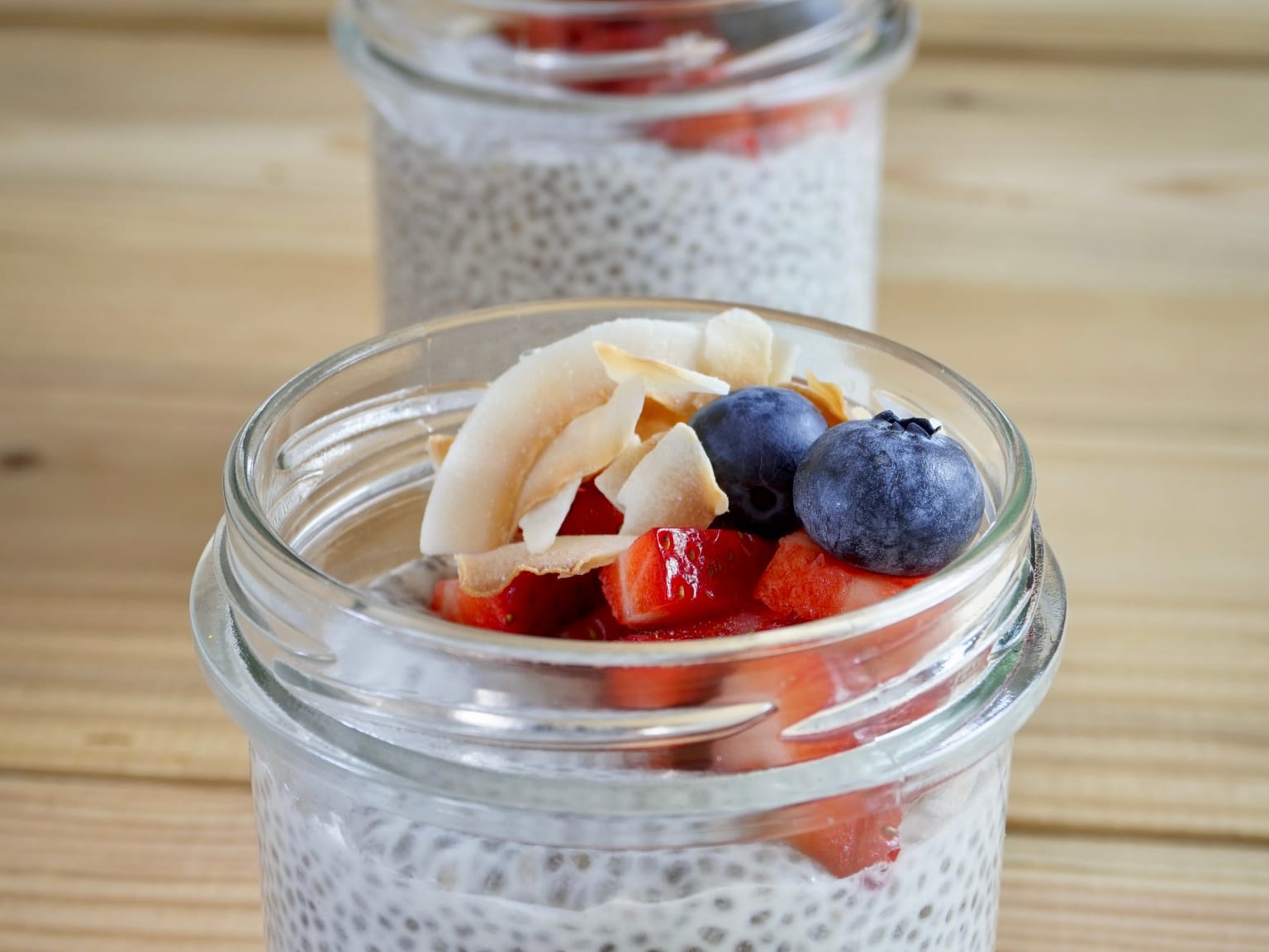 A glass jar of pale chia pudding on a cream surface in natural light, topped with raspberries, a spoonful of Greek yogurt, and chopped walnuts