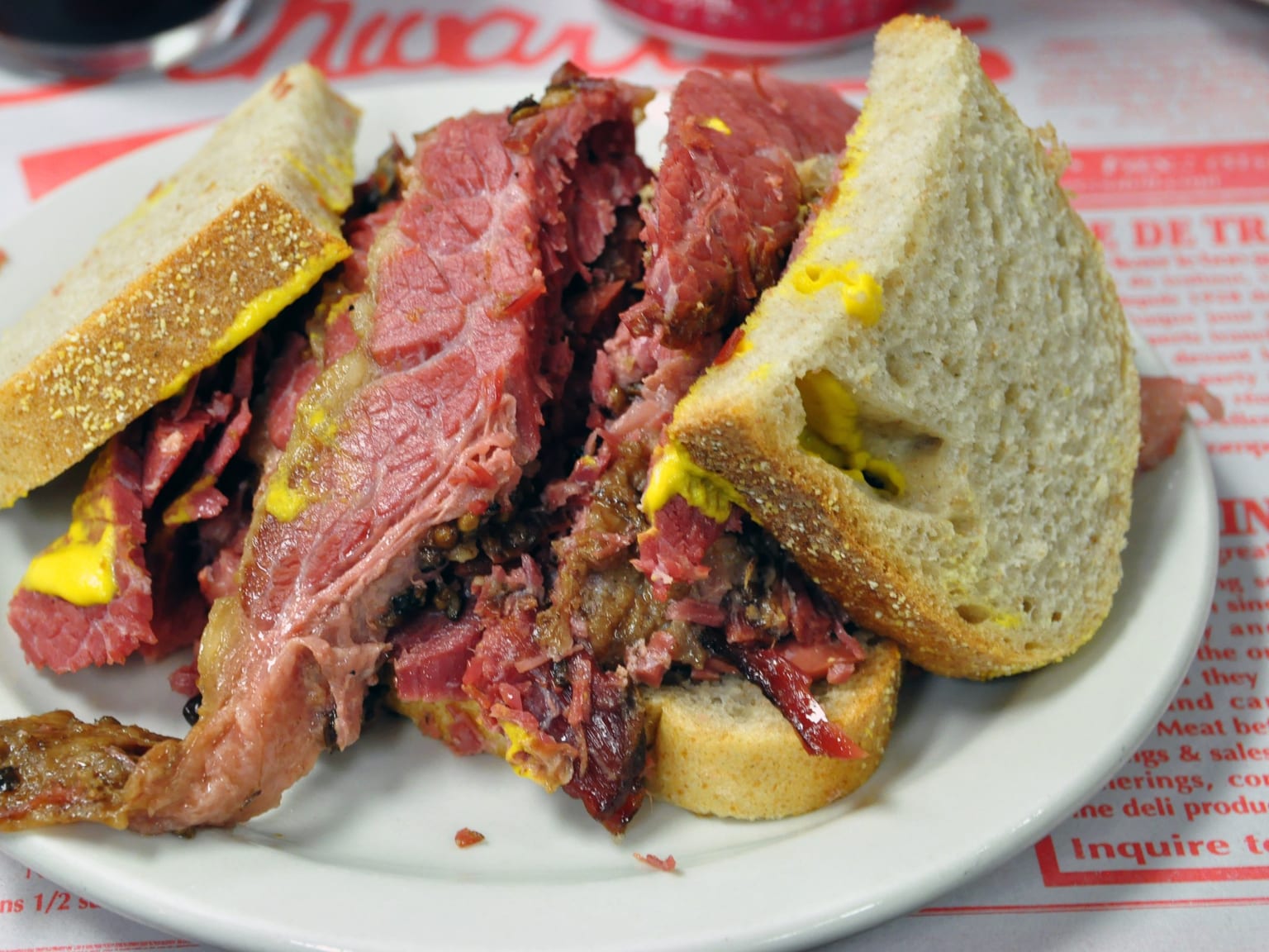 A hand-cut pile of dark peppered Montreal smoked meat between two thin slices of rye bread on a cream plate in natural light, the slices thick and ragged with a visible fat gradient, a smear of yellow mustard and a dill pickle beside it