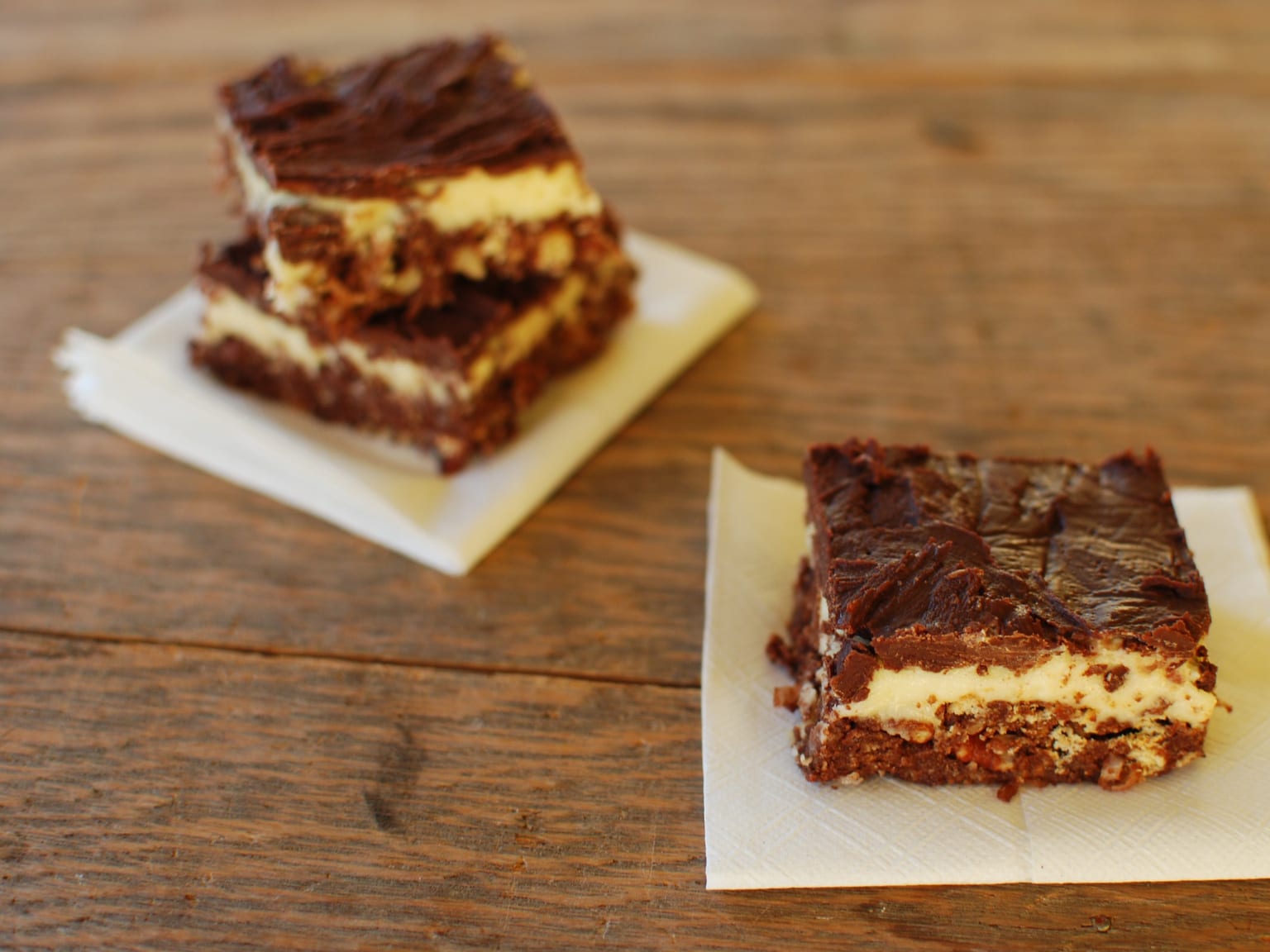 Sixteen Nanaimo bars on a cream ceramic plate in natural light, cut clean so each square shows the dark crumb base, the pale yellow custard layer, and the thin glossy chocolate top
