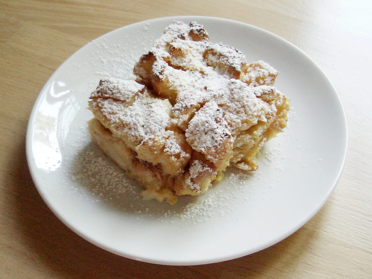 Ofenschlupfer in a cream rectangular baking dish, browned bread edges standing up through set custard with apple slices and toasted almonds, a jug of Vanillesoße beside it in natural light