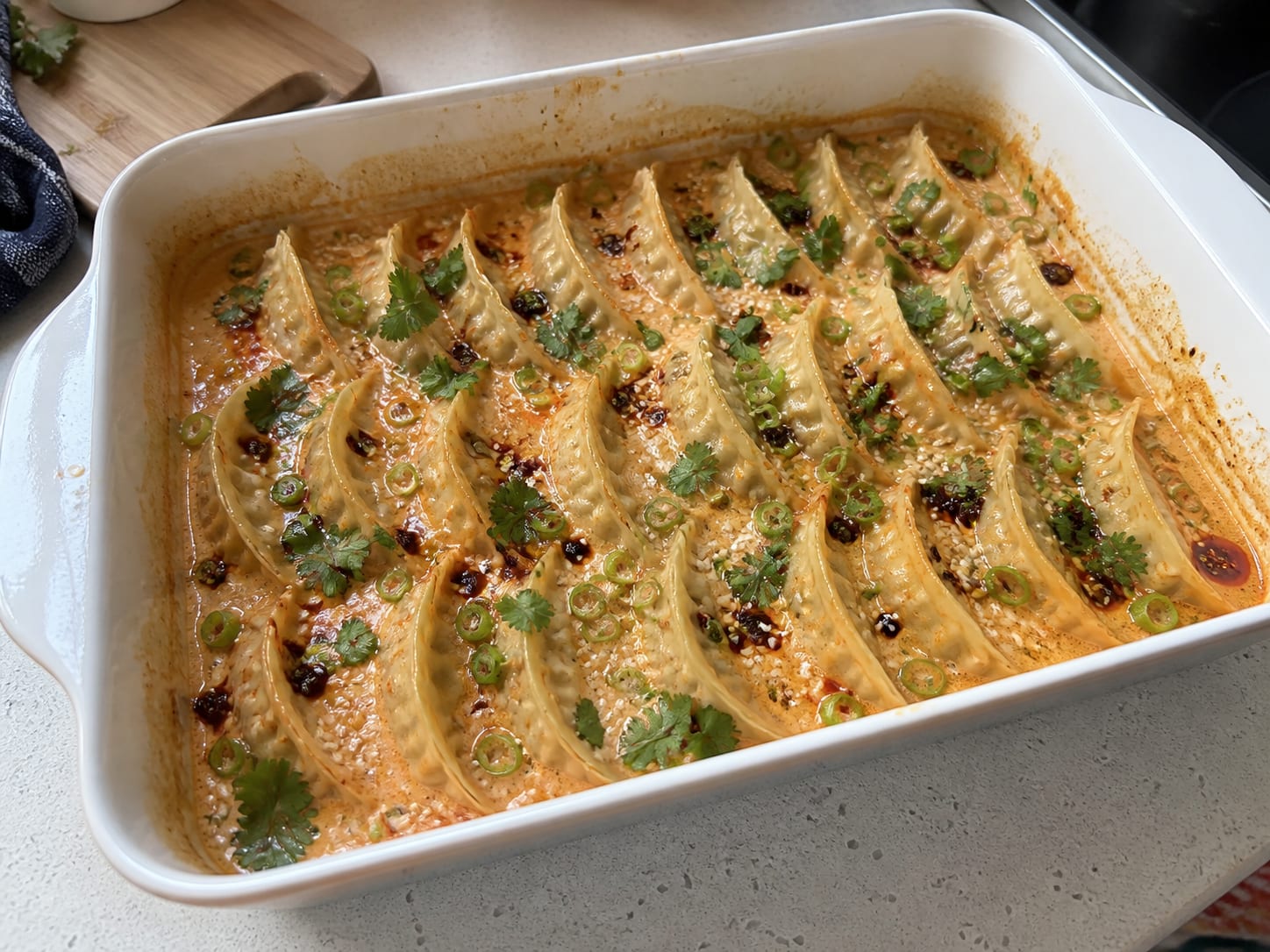 Eine weiße Auflaufform mit Teigtaschen, gebacken in einer cremigen orangefarbenen Curry-Brühe, garniert mit Frühlingszwiebelringen, frischem Koriander, Sesamsamen und knusprigem Chiliöl