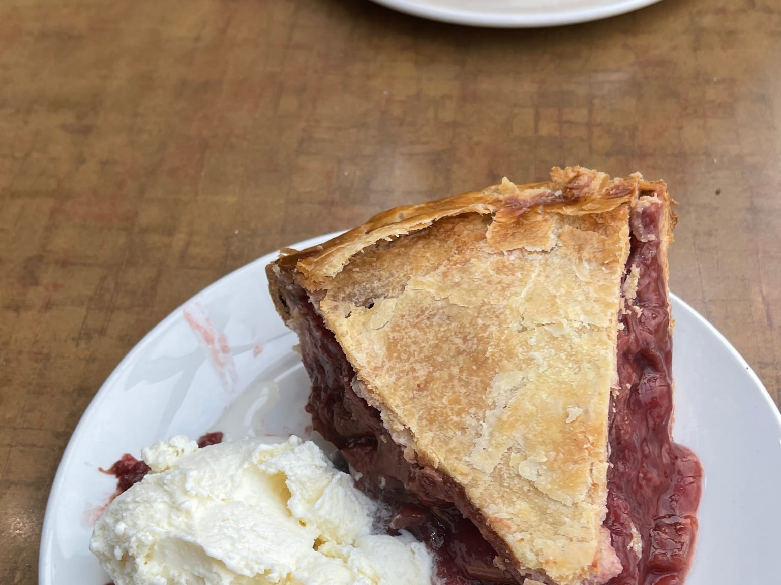 A slice of double-crust plum pie with dark jammy fruit spilling from under a flaky pale-gold lid, with whipped cream beside it