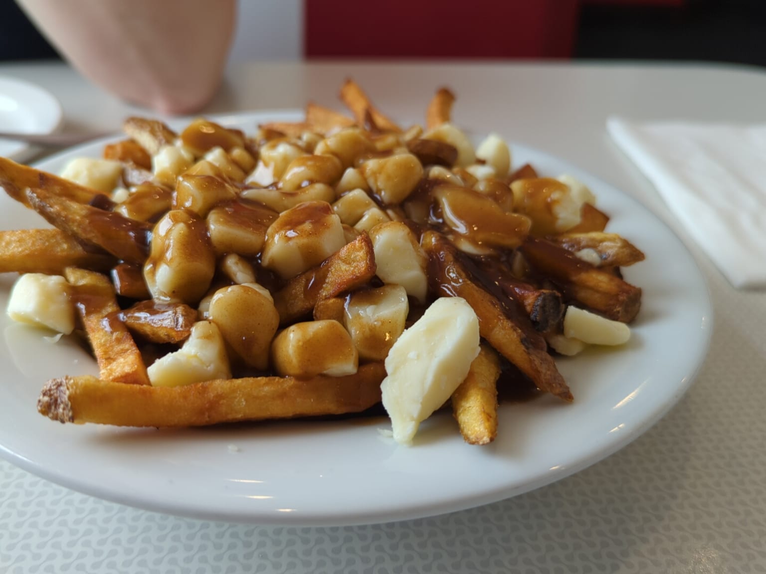 A deep bowl of poutine on a cream table in natural light, thick golden chips under pale lumps of cheese curd half melted at the edges, glossy dark brown sauce poured over the top and running down the sides