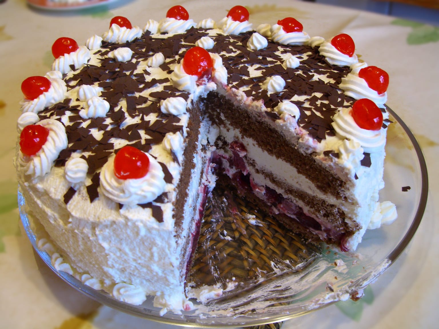 A tall Schwarzwälder Kirschtorte on a cream cake stand, dark chocolate curls over whipped cream, twelve sour cherries around the rim, one slice cut to show the shortcrust base under the cherries and the three soaked sponge layers
