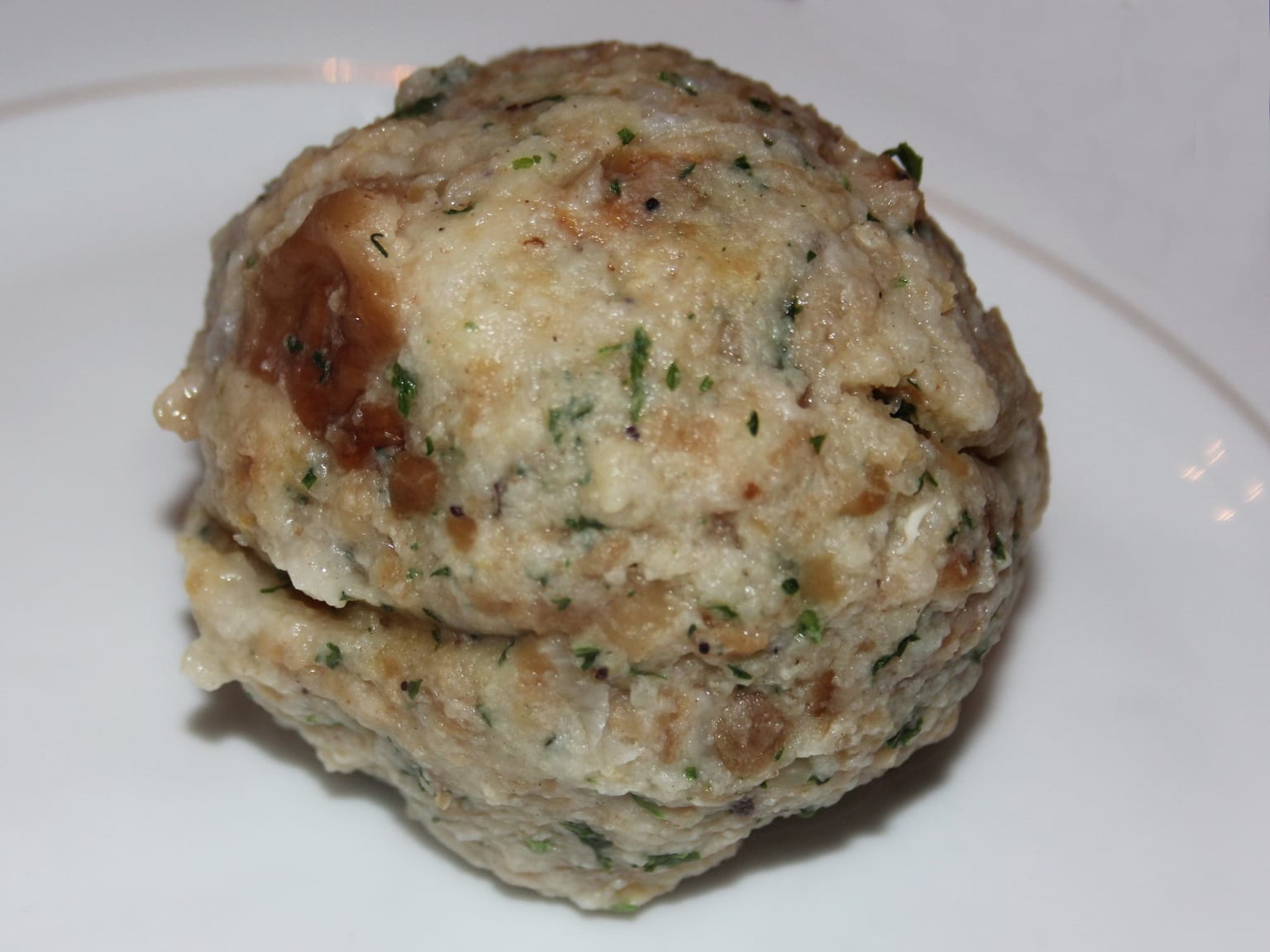 Three pale bread dumplings on a cream plate beside a spoonful of dark gravy, parsley flecks visible in the cut surface