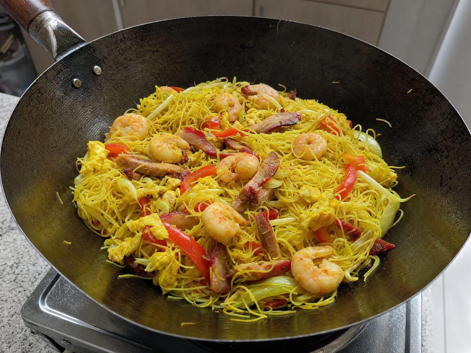 A wok of yellow curry-coated rice vermicelli with prawns, strips of BBQ pork, egg and bean sprouts