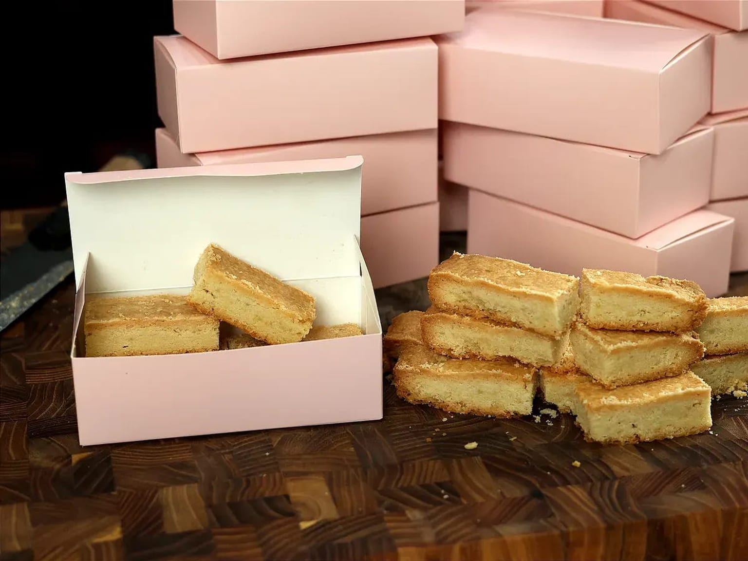 Golden square shortbread biscuits beside an open pink pastry box, with more pink boxes stacked behind