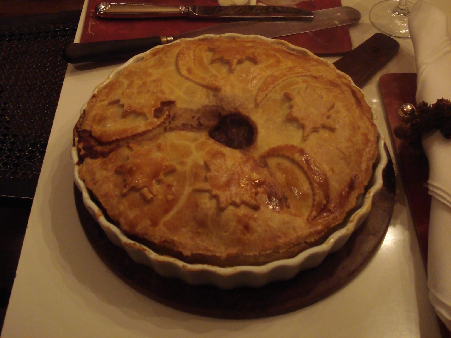 A golden 23 cm tourtière on a cream plate in natural winter light, the top crust glazed and cut with three steam vents, one wedge lifted out to show the dense spiced pork filling