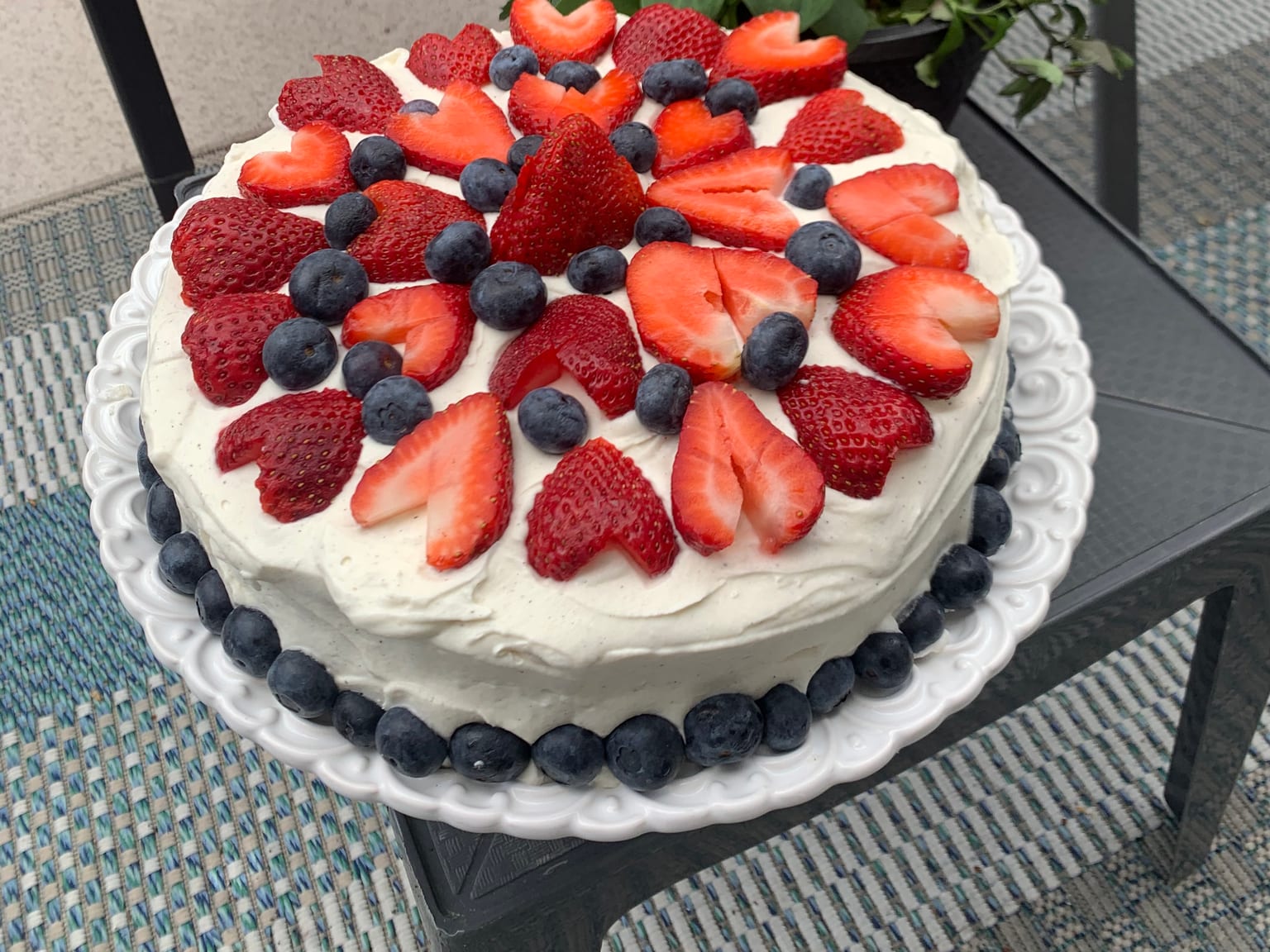 Ein runder Kuchen unter dicker, von Vanillesamen gesprenkelter Creme, obenauf halbierte Erdbeeren und Heidelbeeren, ein Ring aus Heidelbeeren am Rand, auf einer weißen Platte im Freien