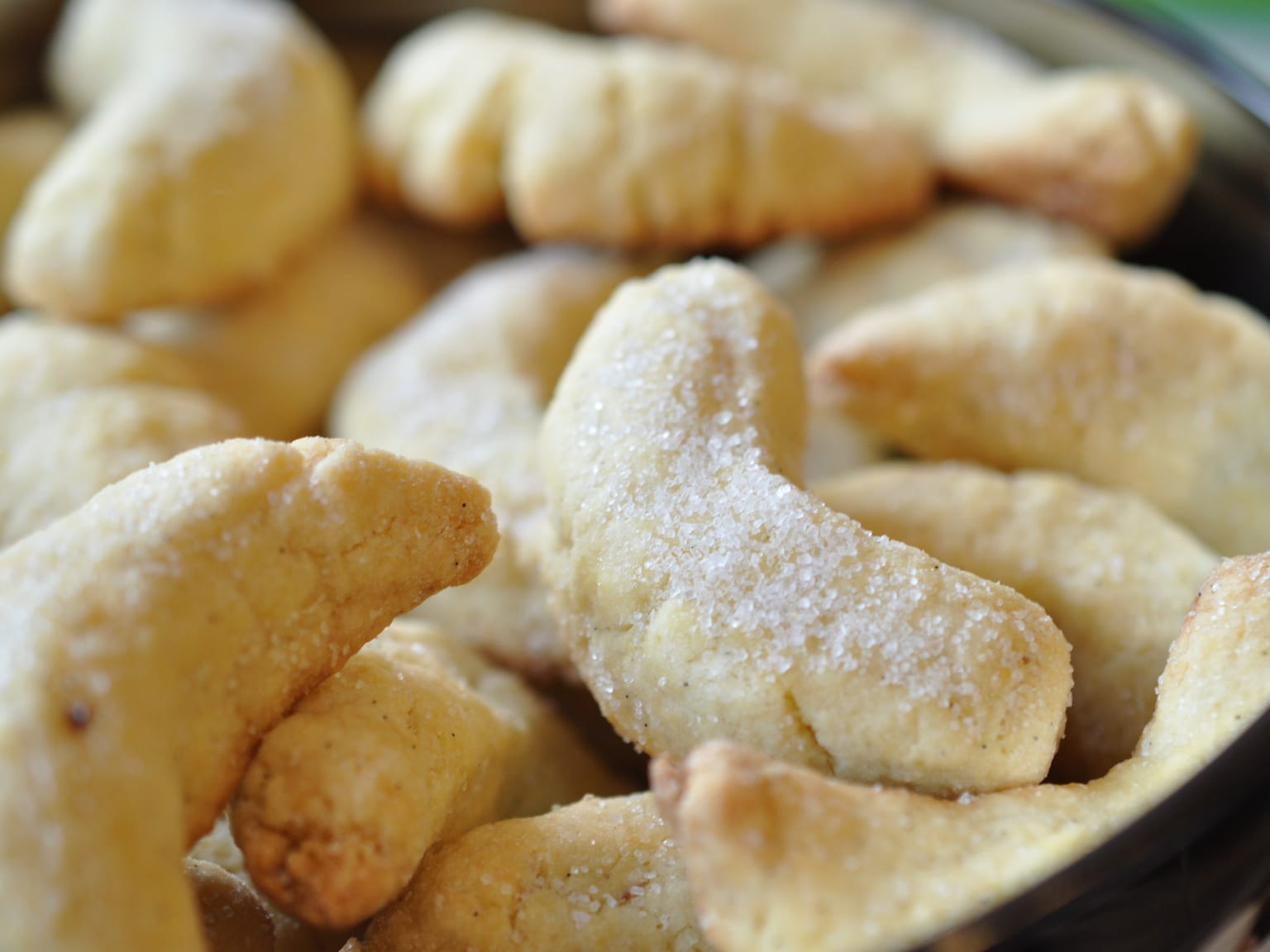 A cream ceramic plate of pale Vanillekipferl heavily dusted with vanilla sugar, natural light from the left, a few loose crescents and a scraped vanilla pod on the linen beside it