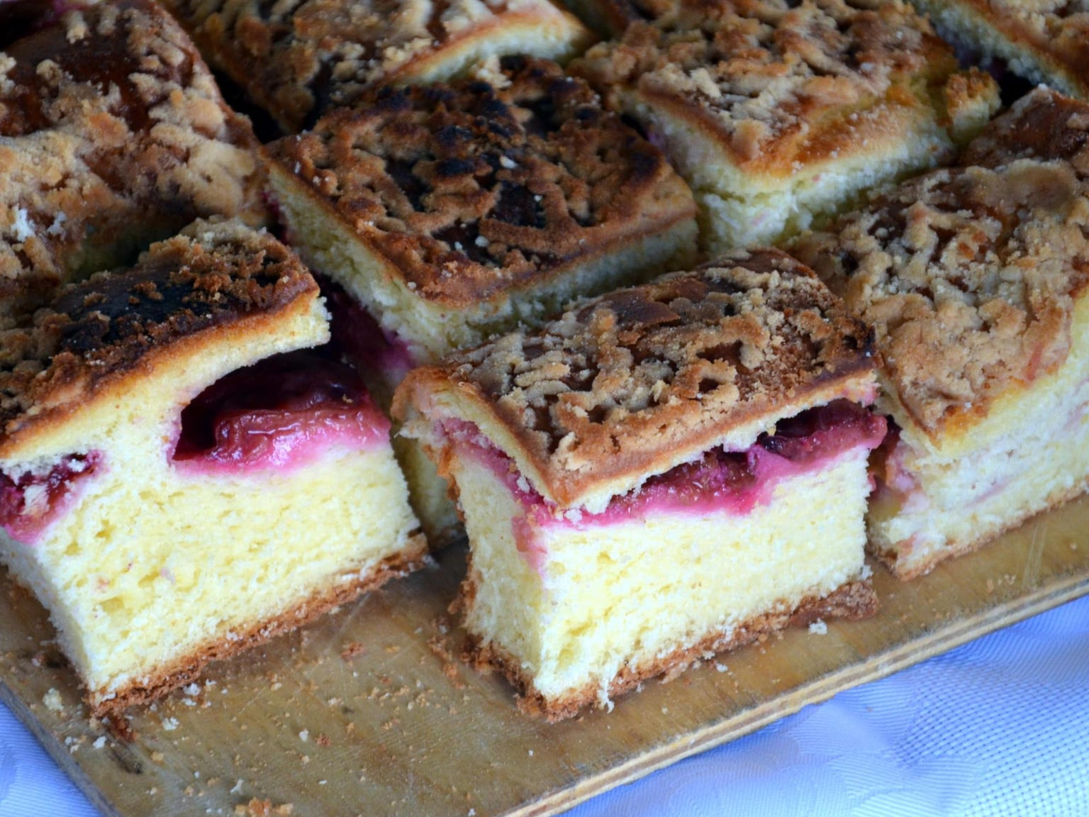 A 30 by 40 cm sheet of Zwetschgenkuchen on a cream cloth, dark blue plum halves packed cut side up in overlapping rows over pale yeast dough, one corner cut away to show the dry golden base, a bowl of whipped cream beside it in natural light