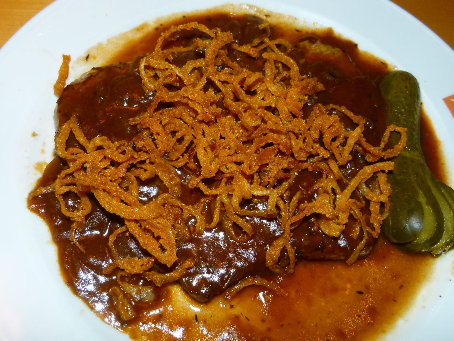 A seared rump steak on a cream plate under dark onion sauce, piled with crisp golden fried onion rings in natural light